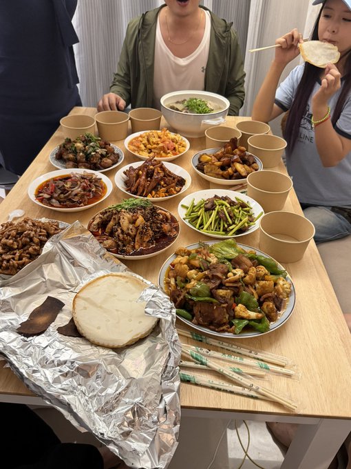A table spread with various Chinese dishes, bowls, chopsticks, and a flatbread on foil, with two people visible at the edges of the frame.