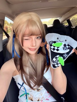 The image features a woman sitting inside a car, holding up a toy pizza that has a face on it. The woman appears to be posing for a photo while holding the toy pizza. The background of the image shows a car interior with a seatbelt visible in front of her.