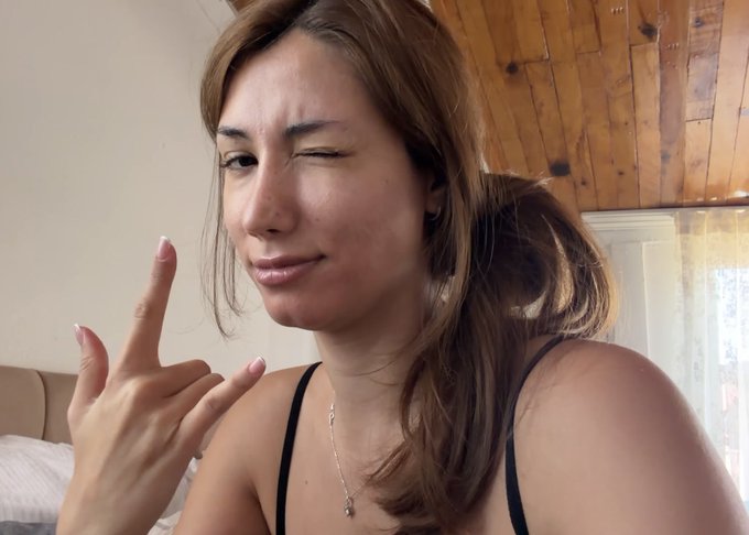 A young woman taking a selfie while winking and making a finger gesture, with light brown hair tied back, wearing a black tank top and necklace in a room with wooden ceiling.