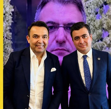 Two men in suits stand smiling together in front of a large purple-tinted portrait of a man wearing glasses, with floral decorations visible in the background. The image appears to be from a formal event or press gathering, though no specific names, text, or identifiers are visible in the image itself to directly connect it to the accompanying post about Murat Ağırel and Tahir Sarıkaya's prosecutorial statement.