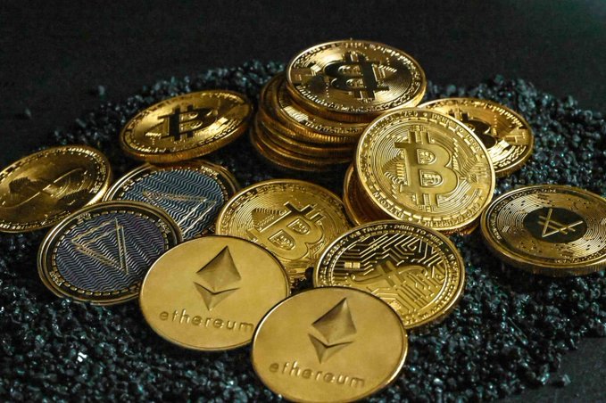 A collection of physical cryptocurrency coins, including gold Bitcoin and Ethereum coins, along with other altcoin tokens, resting on a dark granular surface.