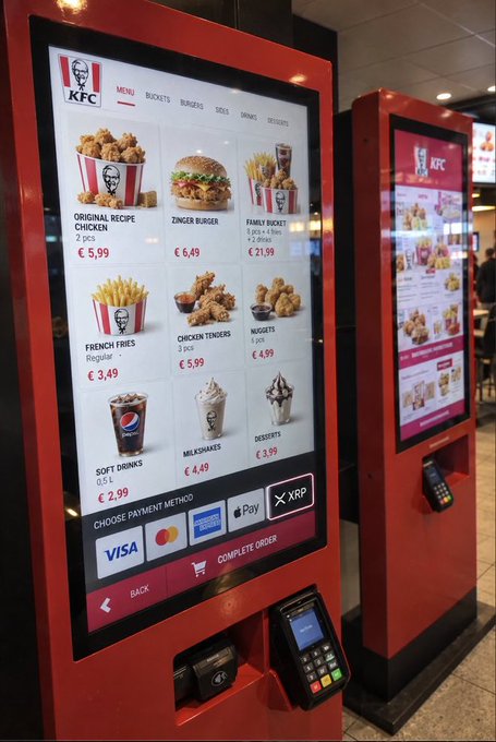 Two red KFC self-ordering kiosks display a digital menu featuring items such as the 2 pcs Original Recipe Chicken (€5.99), Zinger Burger (€6.49), Family Bucket (€21.99), French Fries (€3.49), Chicken Tenders (€5.99), and Milkshakes (€4.49), with payment options for XRP, Apple Pay, and Visa at the bottom.