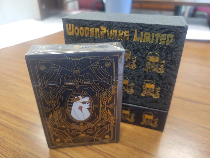 Two decorative playing card boxes from WoodenPunks Limited on a wooden surface - a black box with gold designs featuring a queen card in front, and a textured black box with gold robots behind it.