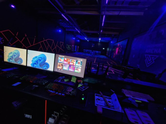 A dimly lit gaming arena with blue and purple neon lighting, featuring a row of gaming PCs with triple monitors, RGB keyboards, and chairs arranged for players, with an 'ARENA' sign visible on the right wall.
