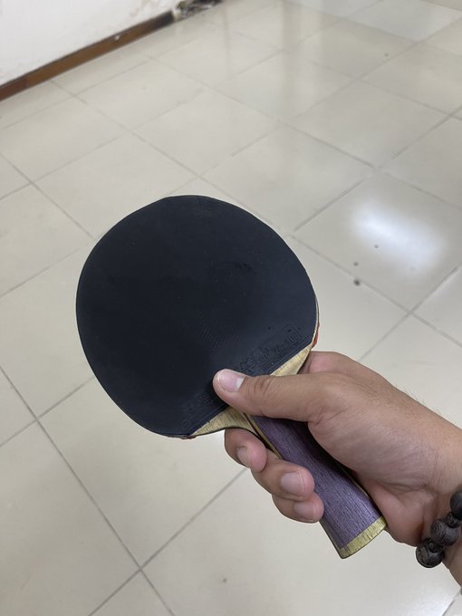 A hand holds a black table tennis paddle with a purple-wooded handle against a backdrop of light-colored tiled flooring.