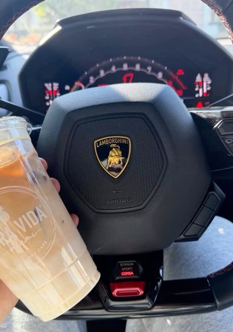 A first-person view from the driver's seat of a Lamborghini with a Corsa driving mode button visible, while a hand holds a clear plastic cup of iced coffee in the foreground.