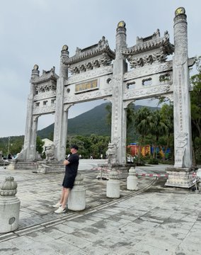A person in dark clothing and shorts stands on a stone plaza in front of an ornate traditional Chinese stone archway with mountains visible in the background.