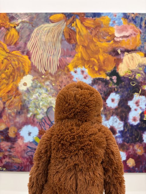 A figure covered in shaggy brown material viewed from behind, standing in front of a large abstract painting filled with warm orange and purple floral shapes.