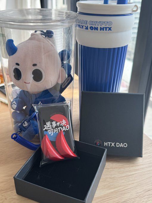 A collection of HTX-branded merchandise including a plush mascot, a blue "Trade Crypto Only on HTX" tumbler, a "遇见未知 问问DAO" (Meet the Unknown, Ask DAO) enamel pin, and an "HTX DAO" gift box, all styled in Huobi Blue. The post pairs Monday with the signature blue Huobi branding, inviting followers to pick their favorite promotional item.