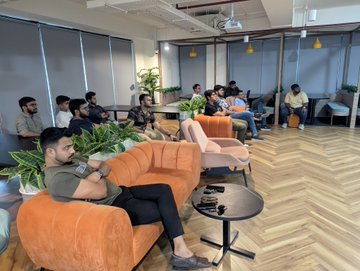 Several people are relaxing in a modern office lounge area with orange couches, while one person appears to be giving a presentation to the group.