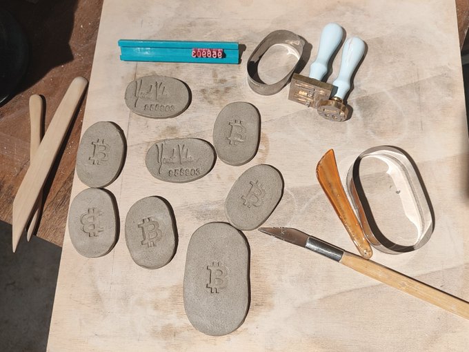 A workbench displaying clay sculptures shaped like Bitcoin coins in various stages of creation, surrounded by crafting tools including carving knives, cookie cutters, and wooden sculpting instruments.