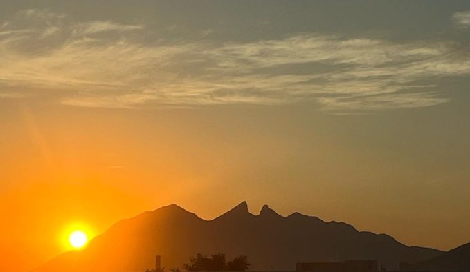 A scenic photograph of a bright orange sunset behind a silhouetted mountain range, with golden-lit clouds stretching across the sky.