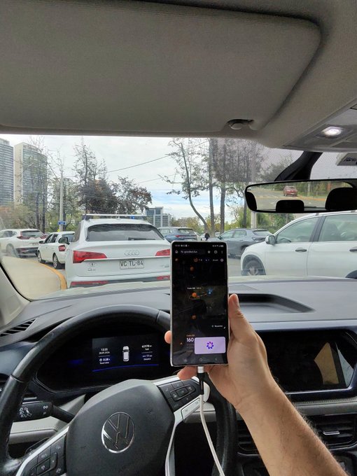 First-person view from inside a Volkswagen vehicle showing a hand holding a smartphone displaying a music player, with the car's dashboard visible and traffic ahead on a city street.