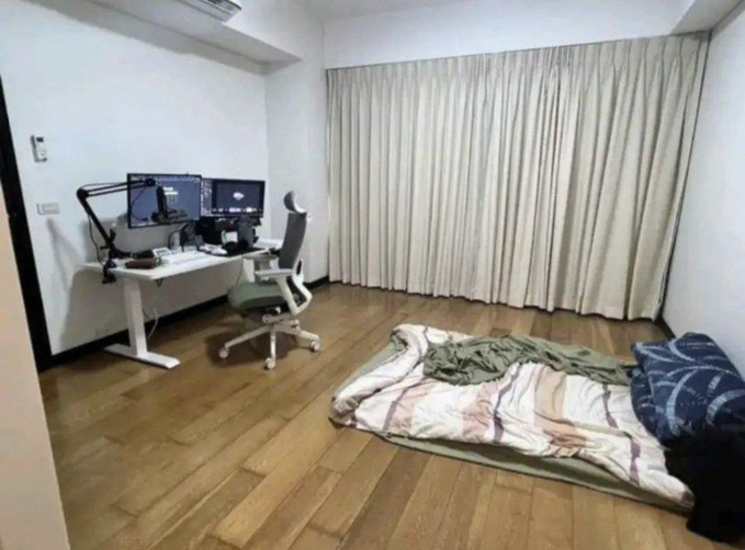 A sparse room with a desk setup featuring dual monitors and an ergonomic chair on one side, while a futon mattress with a pillow and blanket sits directly on the wooden floor on the other side—embodying the "average memecoin trader" lifestyle of dumping rent money into risky bets and living like a college student.
