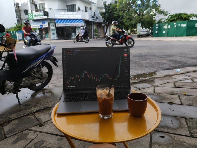 A laptop displaying a stock or crypto trading chart sits on a small yellow table on a street, alongside a glass of iced coffee, with motorbikes passing by in the background.