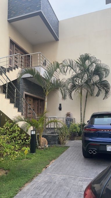 A modern two-story house with an external staircase, palm trees in the yard, and a dark blue Porsche SUV parked in the driveway.