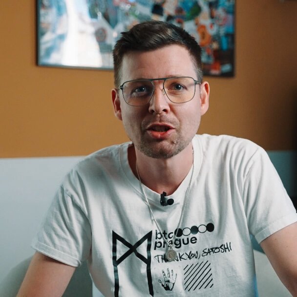 The image features a man wearing glasses, sitting down with his hands resting on his lap. He is dressed in a white t-shirt that has black text printed on it, which reads "MBT Prague". The background of the photo shows an orange wall, and there are two other people visible in the scene - one standing behind the man and another person partially obscured by him.

The post includes a message from someone named "MBT Prague" expressing their thanks for being part of the conversation or event. This message is written in black text on the image.