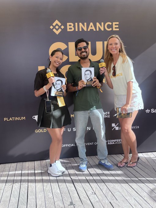 Three people posing at a Binance Club event backdrop, with two women holding microphones and a man in the middle holding a 'Money' magazine, all wearing Binance-branded merchandise.