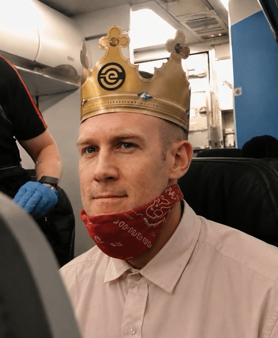 A man wears a Burger King paper crown and a red bandana pulled down beneath his chin while seated on an airplane, with a flight attendant in blue gloves partially visible beside him—visually embodying the "sztorc intensifies" sentiment by combining the meme's iconic crown motif with an exaggerated, deadpan stare.