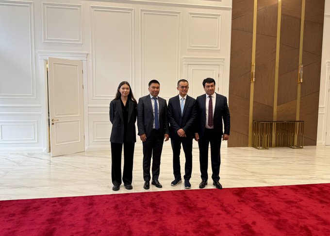 Four people in formal business attire — one woman and three men in suits — stand together posing for a photo in an elegant interior with white paneled walls, a marble floor, and a red carpet.