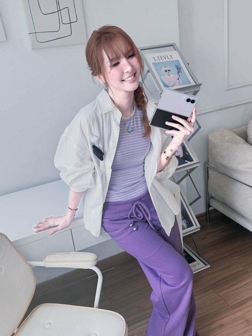 A woman with reddish-brown hair wearing a white jacket, striped shirt, and purple pants, taking a mirror selfie with a phone while holding a book in a room with magazines displayed on a shelf.
