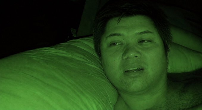 A close-up selfie of a man lying on a pillow in a dark room with a green-tinted lighting effect on the image.