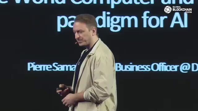 The image shows a man standing on stage at an event, giving a speech or presentation. The man appears to be wearing a tan jacket and has his hands in his pockets while speaking. The background of the image features a large screen displaying text related to the topic of the presentation, which is about AI and its impact on society.