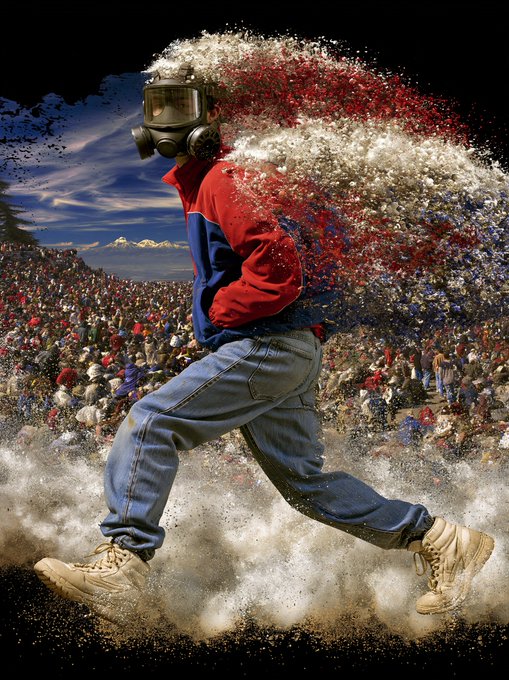 A person wearing a gas mask, red and blue jacket, and jeans appears to be disintegrating into particles while running, set against a backdrop of a large crowd and snowy mountains.