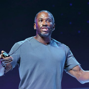 A man in a blue t-shirt stands on stage holding a clicker, appearing to give a presentation or talk.