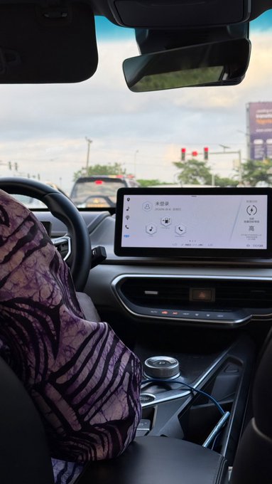 A view from the backseat of a modern car shows a driver in a patterned purple shirt, with the dashboard infotainment screen displaying "2.5GB" and Chinese text, while Chinese city traffic lights glow red in the distance—capturing the moment of moving up through city streets to "unlock" a new level.