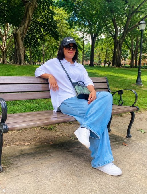 A person wearing a white Nike cap, sunglasses, a white shirt, and baggy blue jeans sits on a wooden park bench.