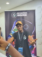 A photo of a young man wearing a black Self Casters Nigeria t-shirt and yellow cap, posing in front of a branded banner while someone takes his picture.