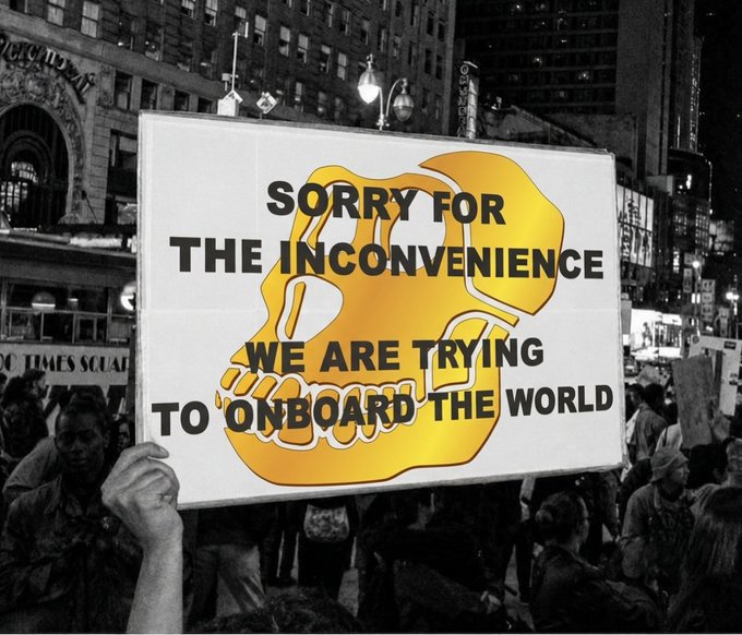 A black-and-white photo of a protest at Times Square with a gold monkey logo overlay and the text 'SORRY FOR THE INCONVENIENCE, WE ARE TRYING TO ONBOARD THE WORLD.'