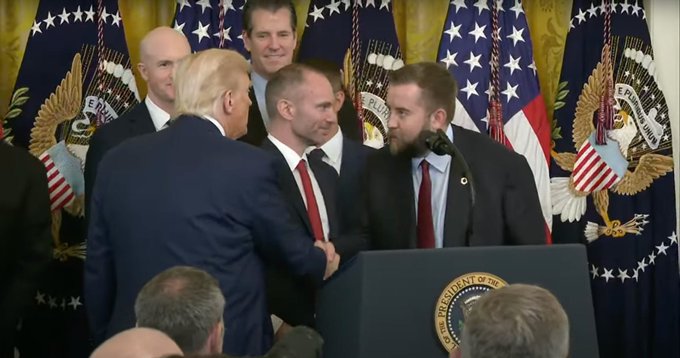 President Donald Trump shakes hands with a man at a podium, while another bearded man speaks at the microphone, with multiple American flags visible in the background.