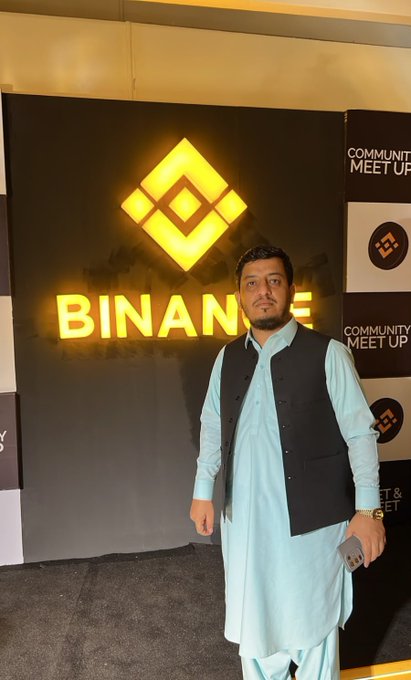 A man wearing a light blue traditional outfit with a black vest stands in front of an illuminated Binance logo at what appears to be a community meetup event.