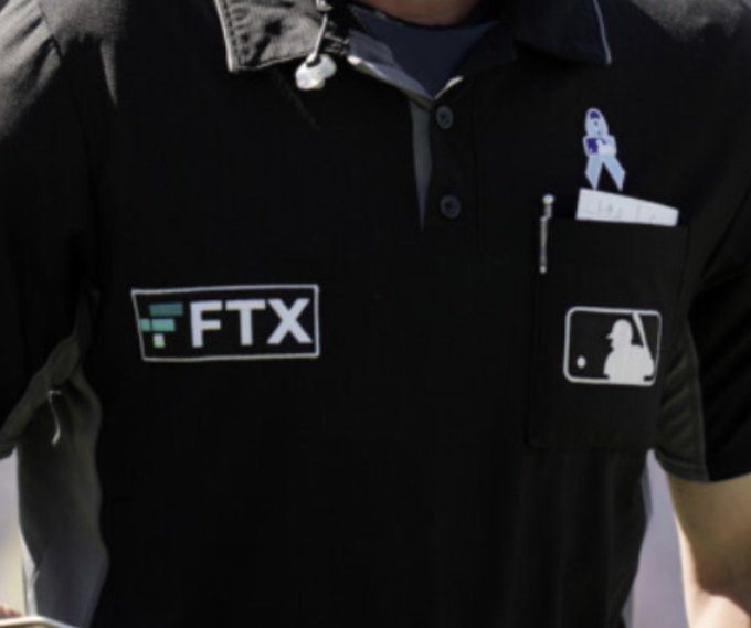 An umpire wears a black MLB shirt featuring an FTX sponsor patch, along with a prostate cancer awareness ribbon pin.
