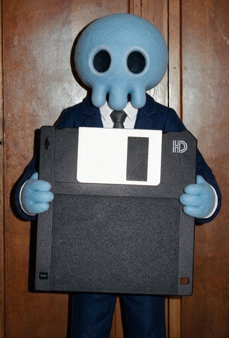 Photograph of a person in a business suit wearing a blue octopus/squid mask, holding a large floppy disk costume piece.