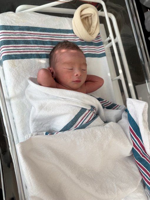 A newborn baby sleeps peacefully in a hospital bassinet, swaddled in white and striped blankets. The image illustrates the post's announcement that the author will be distracted during XYZ's major earnings due to the arrival of their second child.