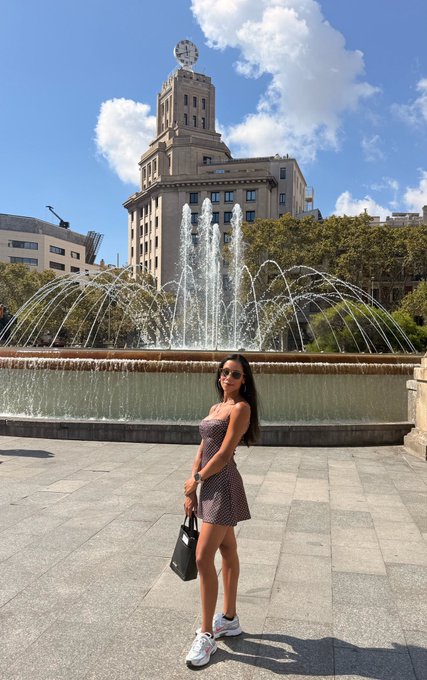 A woman in a polka-dot dress and sunglasses poses in front of a large fountain and historic clock tower on a sunny day.