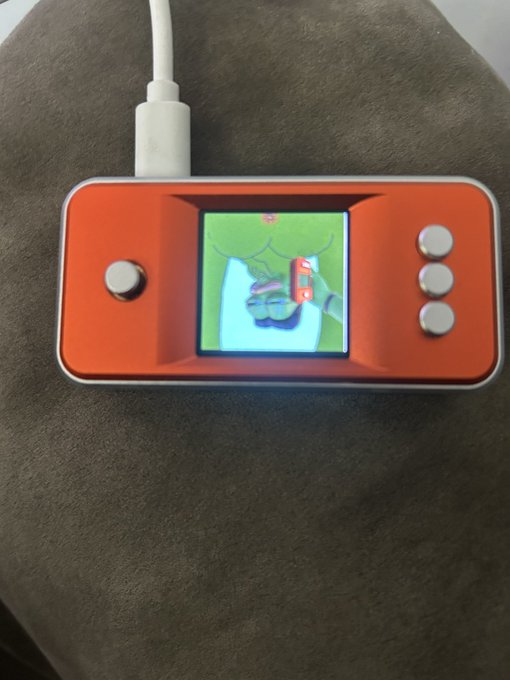 A red handheld retro gaming device connected to a white USB cable, displaying a colorful game on its small screen, resting on a brown textured surface.