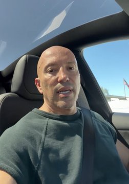 A selfie video of a bald man in a dark green t-shirt sitting inside a car during the daytime.