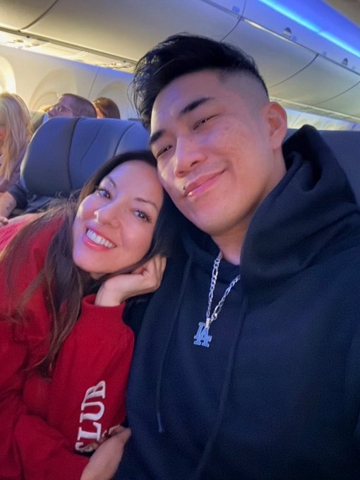 A selfie taken on an airplane shows a smiling woman in a red sweatshirt leaning against a man wearing a black hoodie and a silver 'LA' chain necklace.