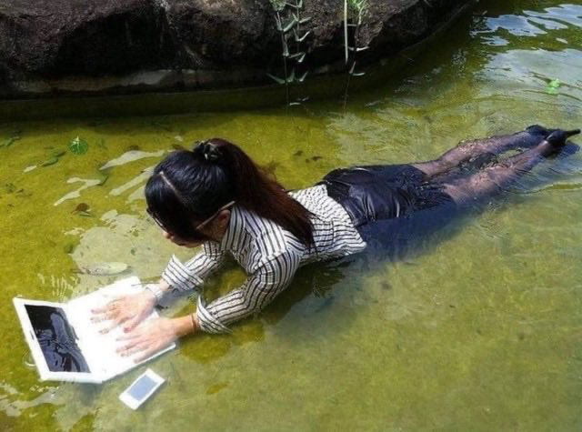 A young woman lies in a shallow pool of water, reading a book placed on the surface, with a smartphone nearby.