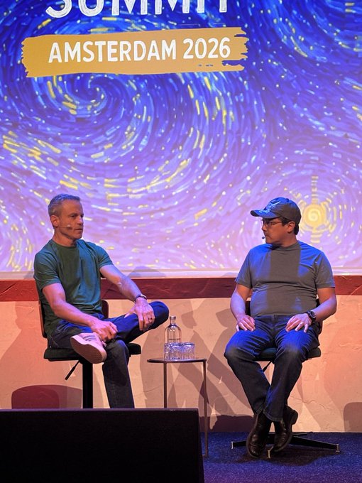 A man wearing a hat sits next to another man who is not wearing one, both sitting on chairs at a stage with a large screen behind them that displays a painting of a spiral galaxy. The background also features a banner that reads "AMSTERDAM 2026".