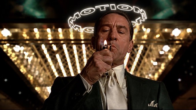 A man in a dark suit lighting a cigarette beneath a glowing 'MOTION' neon sign in an ornate setting.
