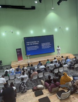 A speaker presents "THE ECONOMIC OS FOR THE INTERNET" on a large screen to an attentive audience seated in a modern conference hall, with the on-stage talk aligning with the post's reference to real builders gathering around the @arc network.