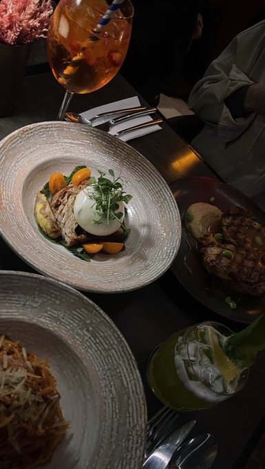 An overhead view of a dimly lit restaurant table featuring plated dishes, an aperol spritz cocktail, and a green drink with lime.