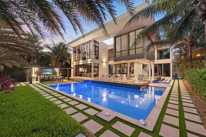 A modern luxury home with large windows, palm trees, and an illuminated blue swimming pool at dusk.