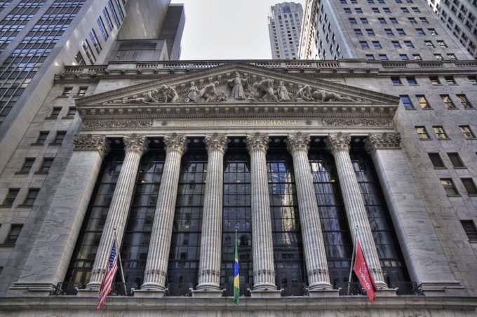A low-angle photograph of the New York Stock Exchange building, featuring its iconic classical columns, sculpted pediment, and three flags (American, green/yellow, and red) at the entrance, with 'NEW YORK STOCK EXCHANGE' inscribed above the columns.