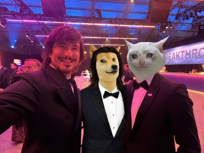 Robinhood CEO Vlad Tenev poses for a selfie with the iconic Robinhood Chain meme mascots, $Cashcat (right) and $VladDog (center), at a "BREAKTHRO" event. The image captures the playful intersection of the meme culture that Tenev himself has actively engaged with on social media, with both characters dressed in formal attire alongside the smiling CEO.